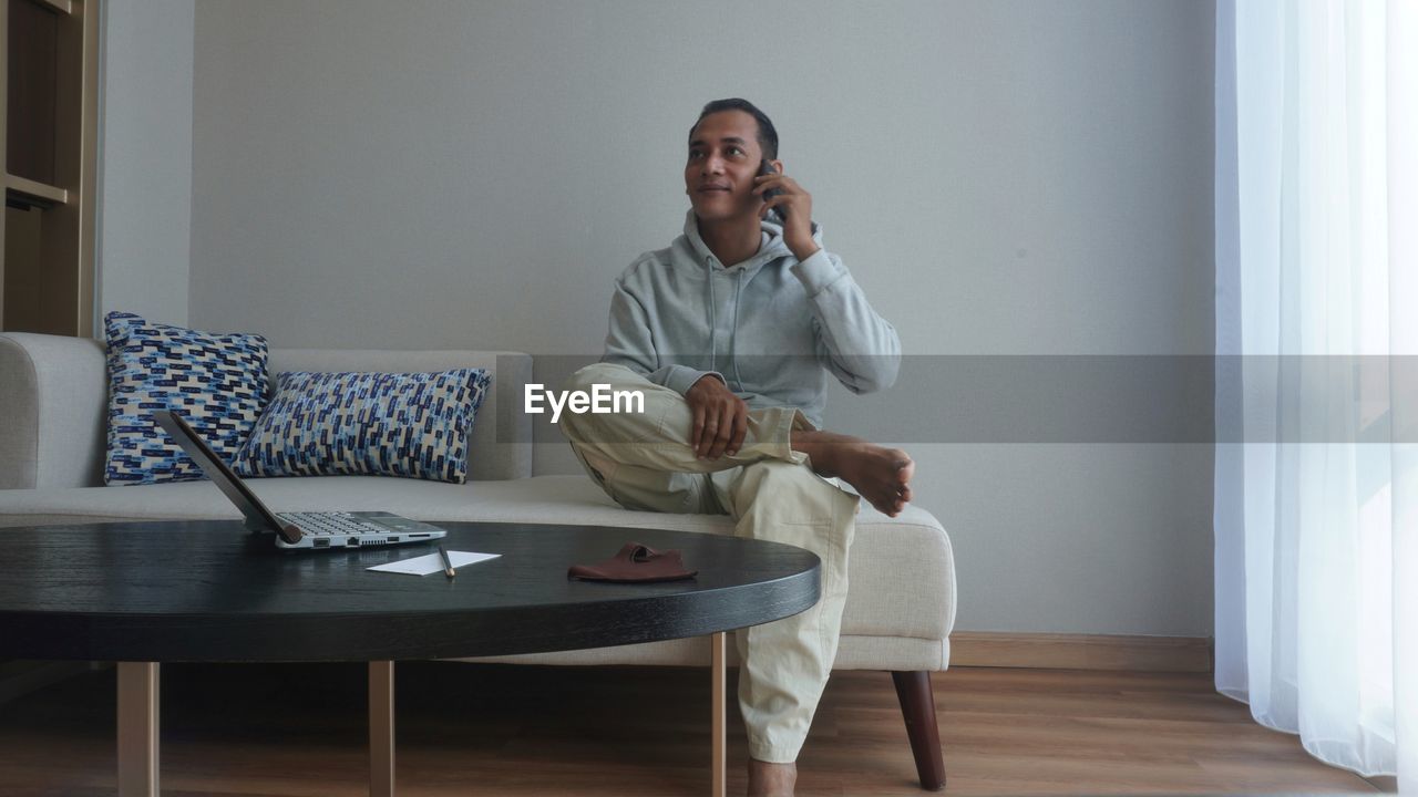 one person, adult, indoors, technology, sitting, wireless technology, communication, telephone, domestic room, home interior, table, furniture, using phone, full length, domestic life, lifestyles, mobile phone, coffee table, clothing, casual clothing, living room, smiling, relaxation, men, person, happiness, emotion, front view, looking, chair, talking, mature adult, room, listening, leisure activity, seat, holding, interior design, portability, sofa, hardwood floor, enjoyment, window, portable information device