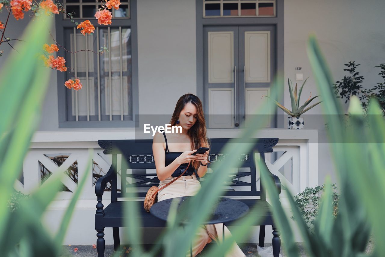 Young woman using smart phone while sitting on chair against house