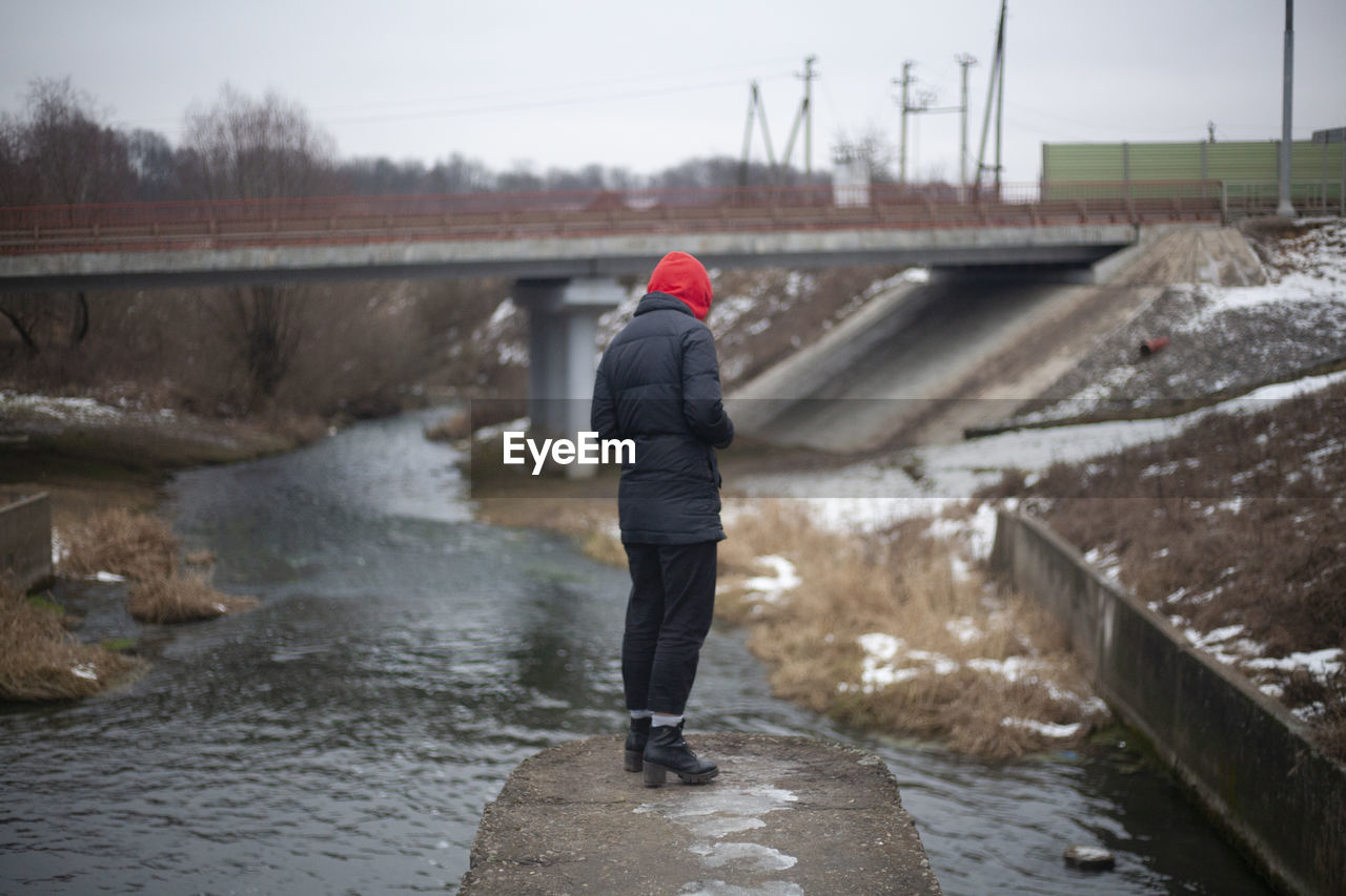 REAR VIEW OF MAN STANDING ON RIVERBANK DURING WINTER