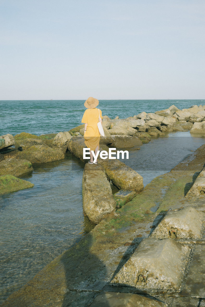 water, sea, shore, rock, horizon over water, one person, sky, horizon, coast, beach, ocean, land, nature, full length, cliff, beauty in nature, vacation, body of water, rear view, scenics - nature, leisure activity, terrain, day, bay, lifestyles, tranquility, standing, clear sky, tranquil scene, women, adult, casual clothing, sand, holiday, outdoors, trip, idyllic, sunlight, wave, non-urban scene
