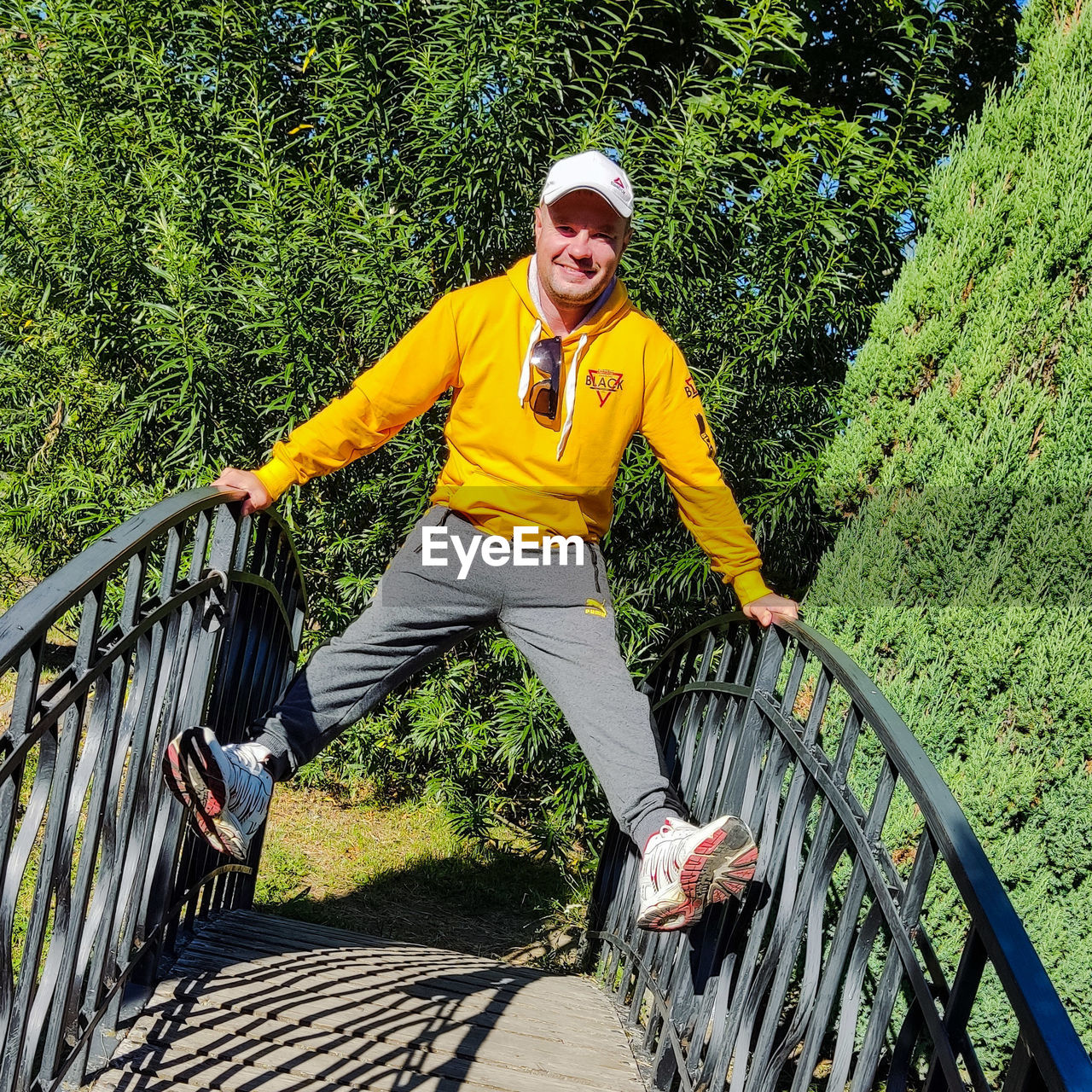 one person, leisure activity, adult, lifestyles, sports, full length, smiling, nature, day, plant, men, front view, happiness, portrait, emotion, looking at camera, railing, casual clothing, outdoors, sunlight, exercising, green, clothing, tree, sports clothing, fun, yellow, vitality, activity, shadow, young adult, sports equipment