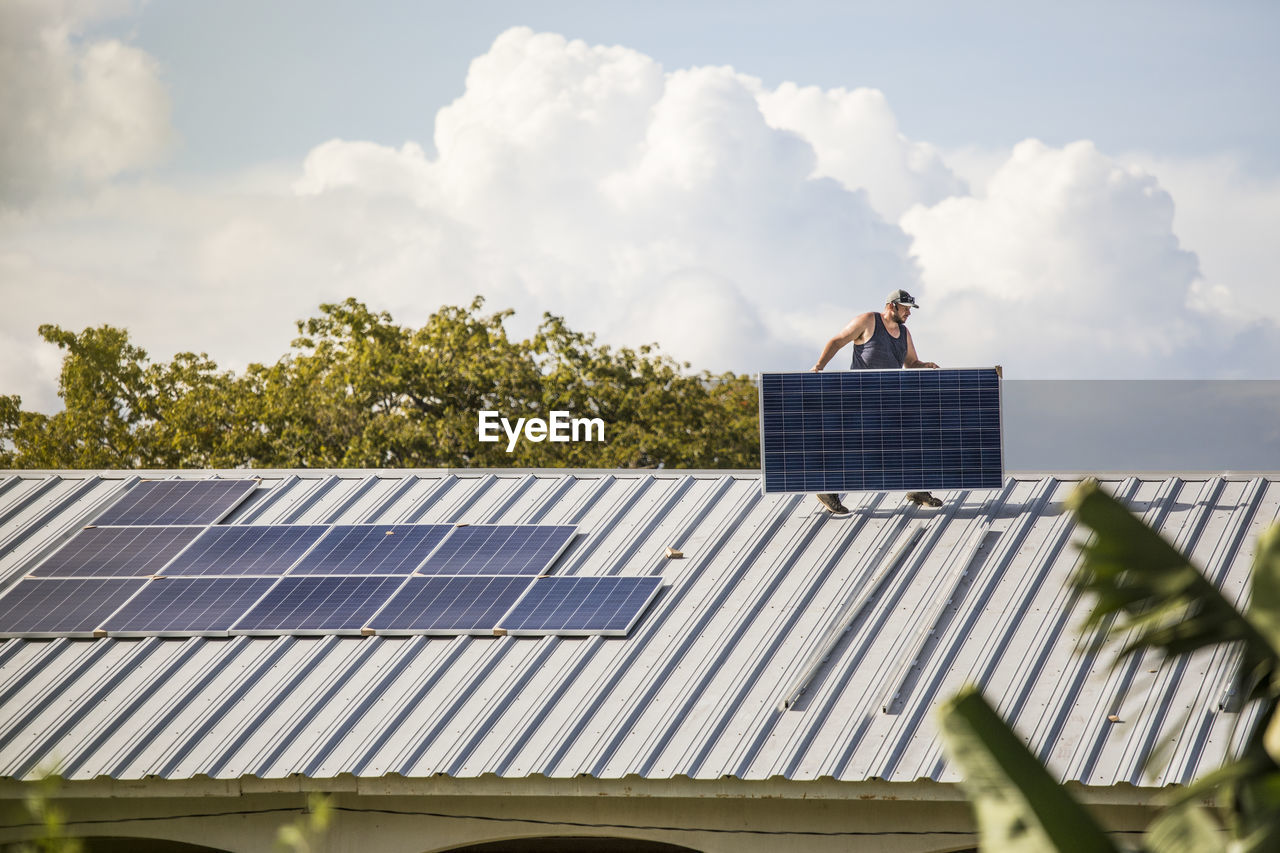 Construction worker carries solar panel across roof during install.