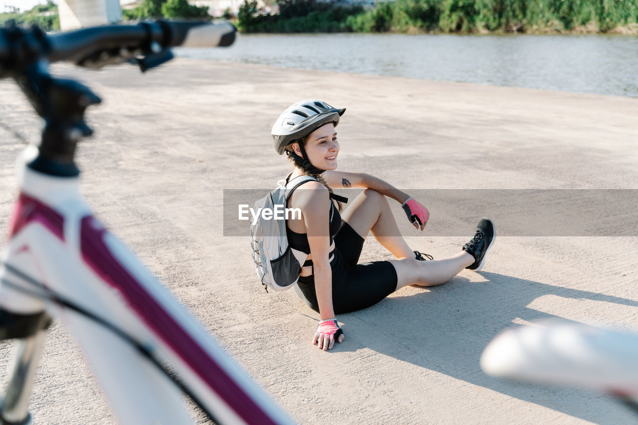 High angle side view of happy young woman in sportswear and helmet with backpack sitting on paved ground and resting after riding bike in summer day