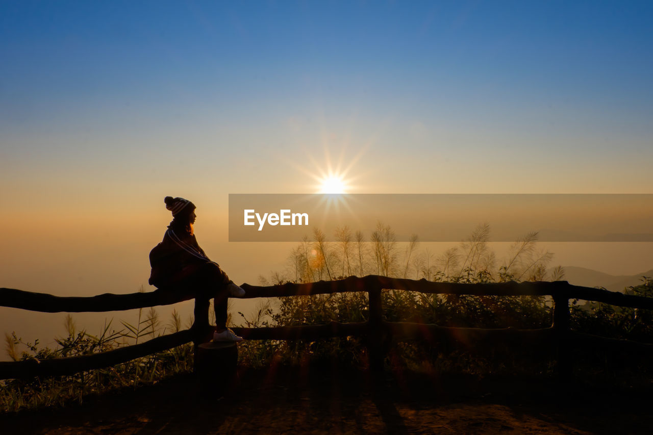 Silhouette woman sitting on fence against bright sun during sunrise