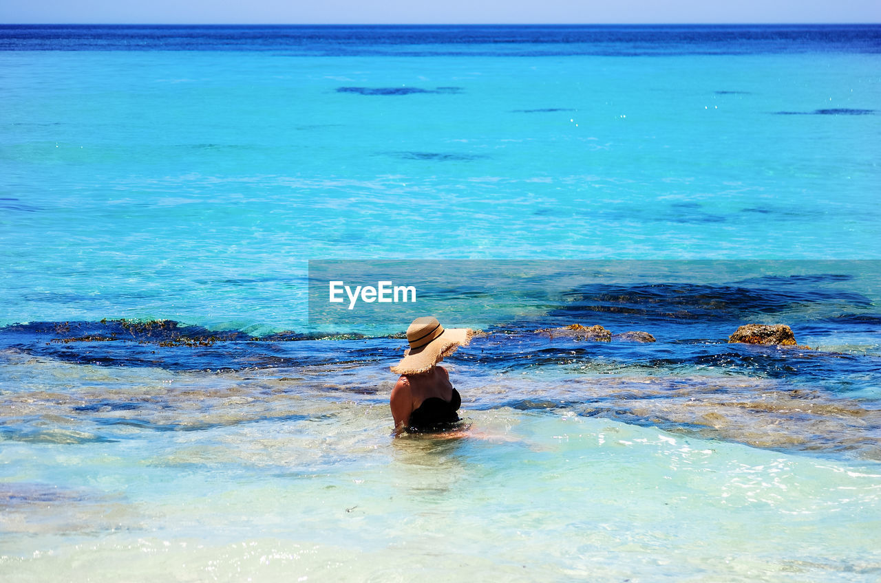 Young woman with straw hat sitting in shallow water of turquoise colored sea