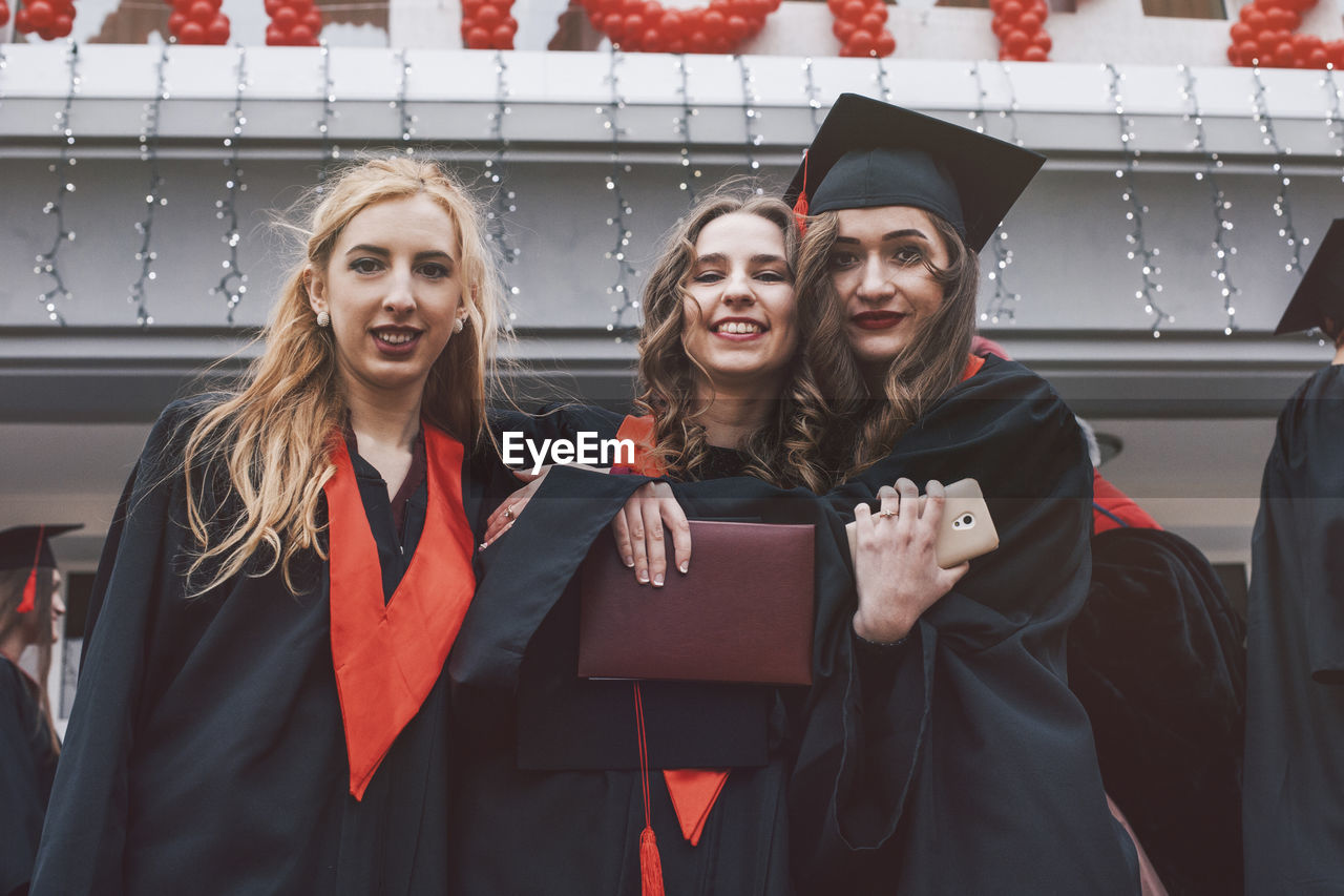 Low angle portrait of happy female students wearing graduation gowns against building