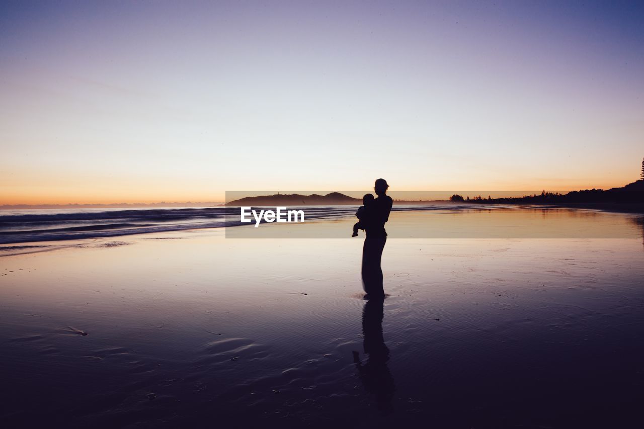 Silhouette mother with her baby at beach against clear sky during sunset