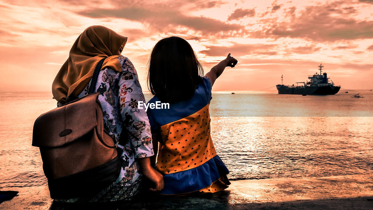 Girl pointing while sitting by mother at beach against sky during sunset