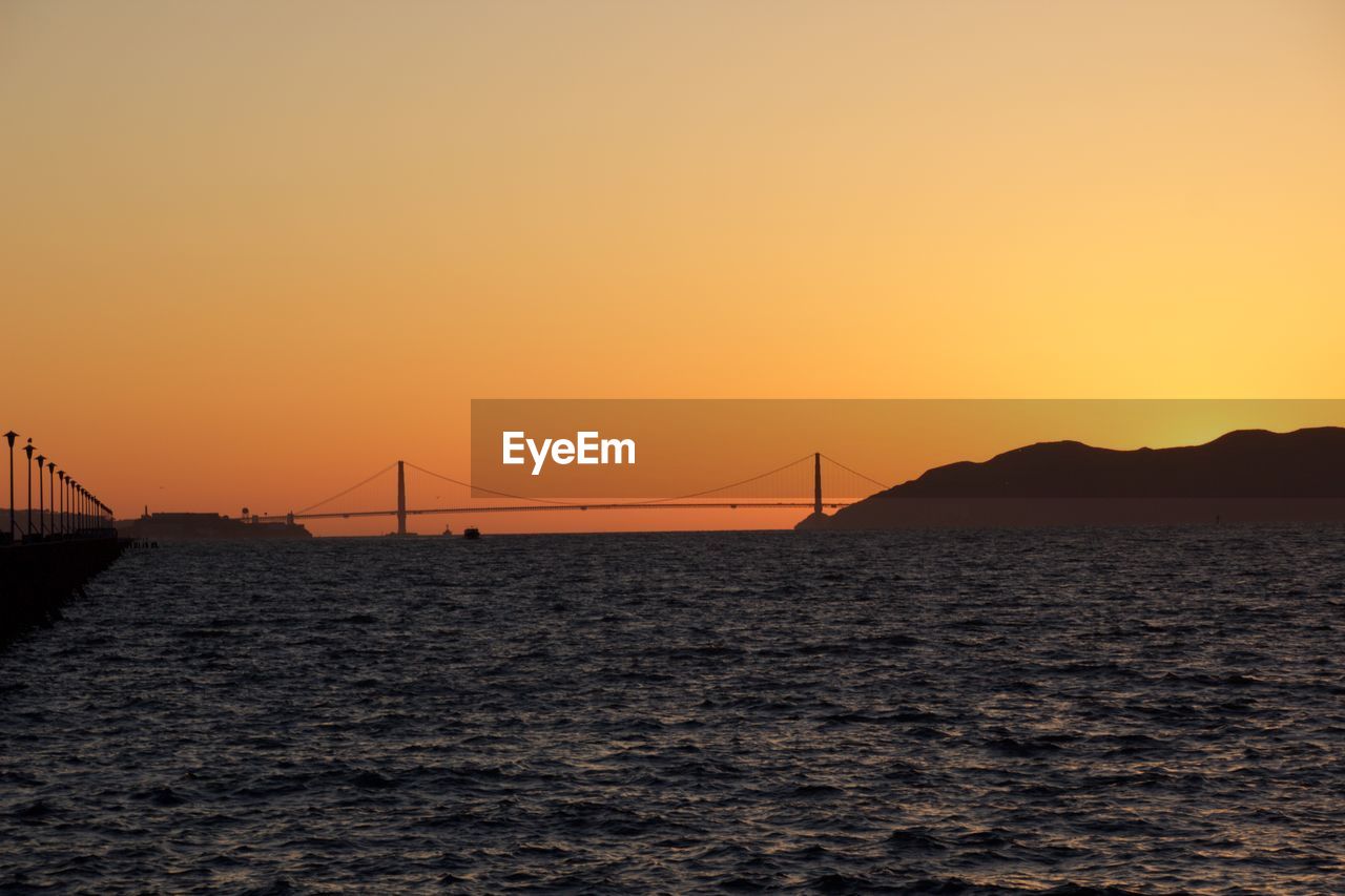 Silhouette bridge over sea against orange sky