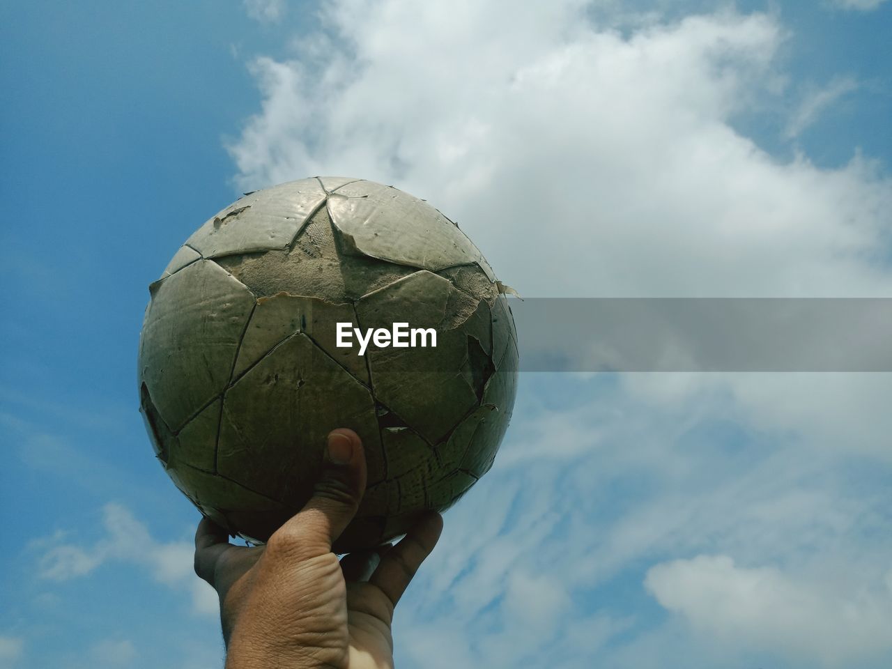 Cropped hand holding old soccer ball against sky