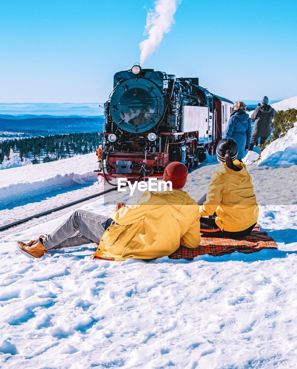 Couple sitting on snow covered land at railroad station against clear blue sky