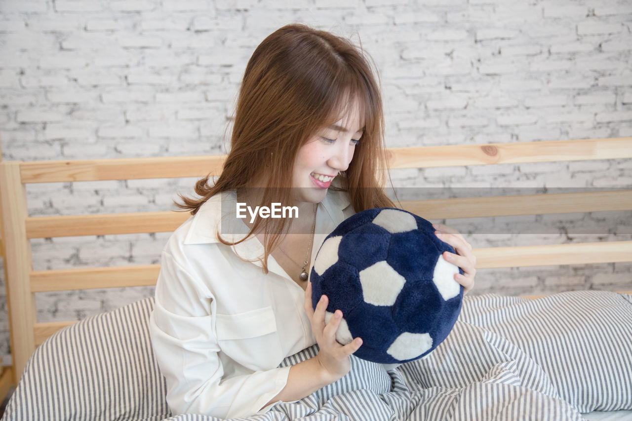 Smiling young woman holding soccer ball while sitting on bed at home