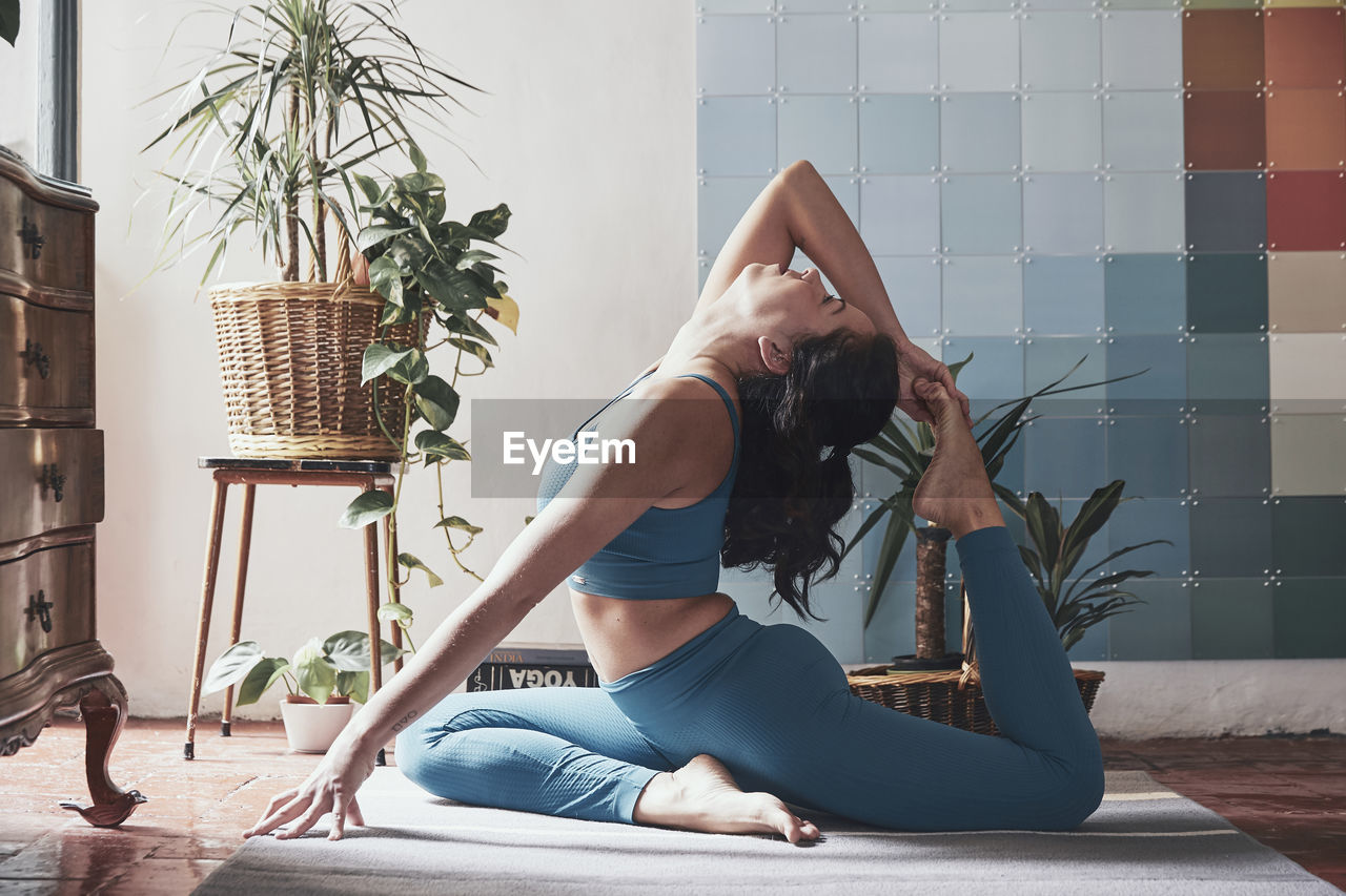 Side view of woman doing yoga while sitting on floor by potted plants