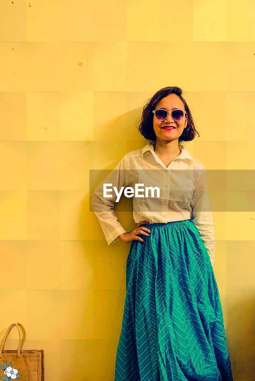 Young woman wearing sunglasses standing against yellow wall in an indo western attire