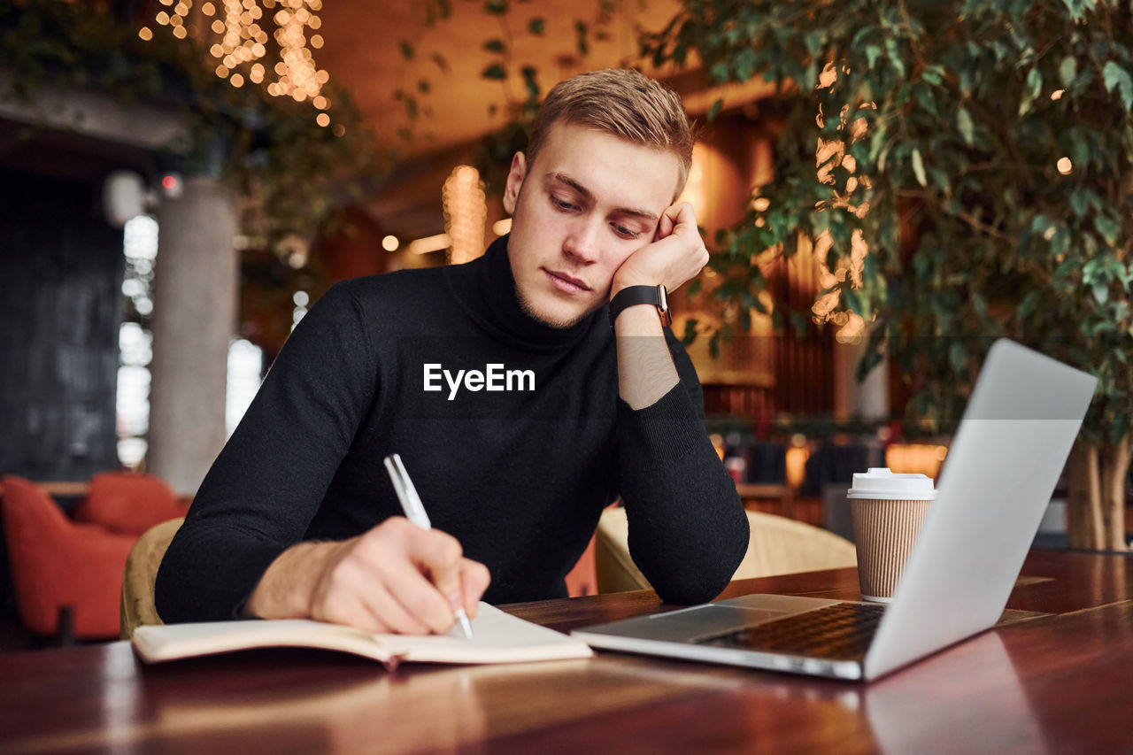 Successful young guy sits in cafe by table with his laptop on it.