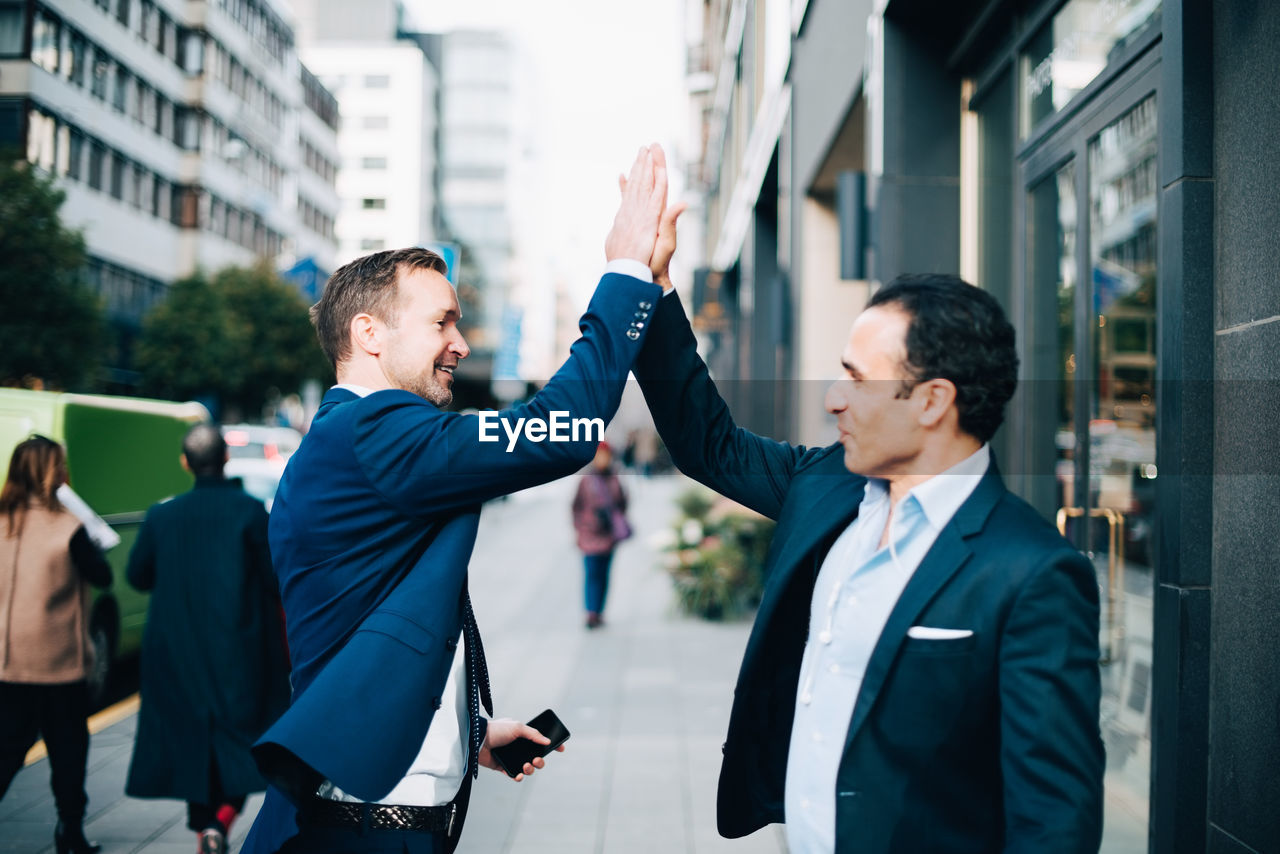 Happy mature male business coworkers giving high-five while standing on sidewalk in city