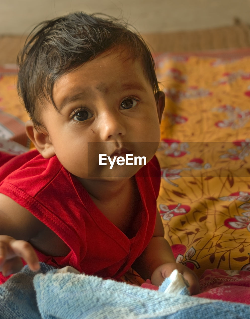 Cute indian baby lying on bed sheet over a mat looking at the camera.