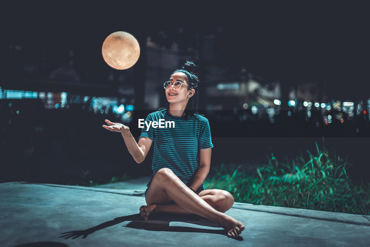 Smiling woman looking at artificial moon while sitting on footpath at night