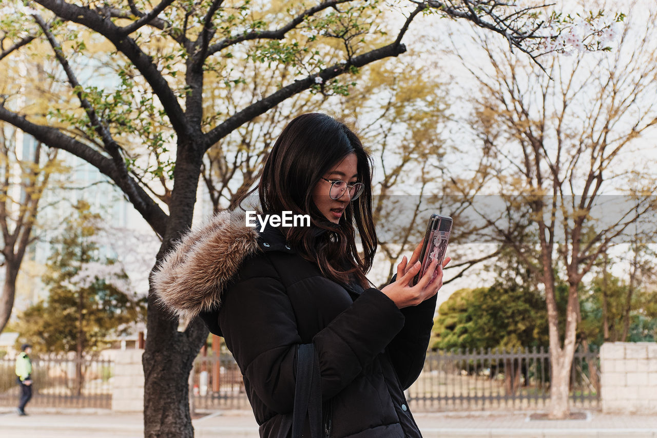 YOUNG WOMAN USING SMART PHONE WHILE STANDING ON TREE