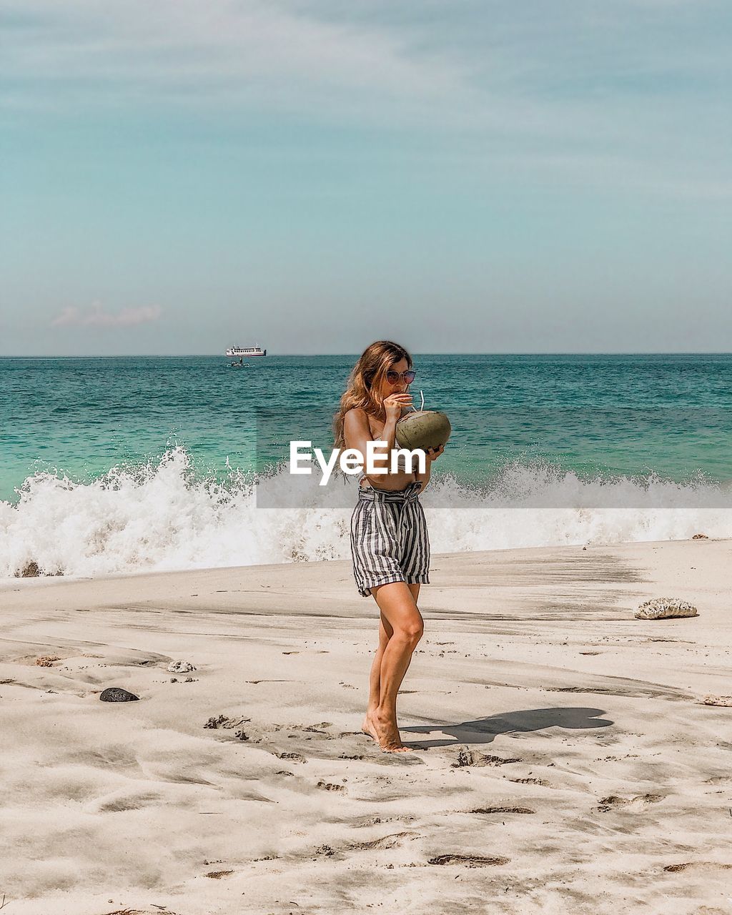 Full length of woman drinking coconut water while standing at beach against sky