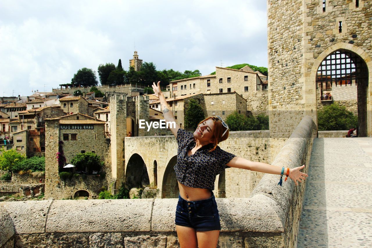 Cheerful young woman with arms outstretched standing against buildings in city