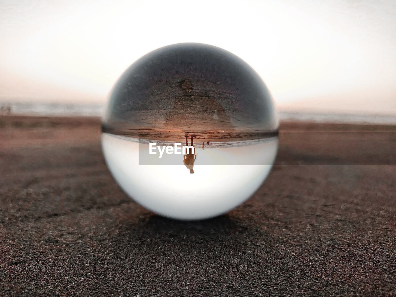 Reflection of man seen in crystal ball at beach against sky during sunset