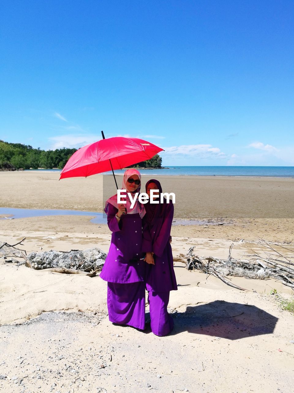 Woman with friend in traditional clothing holding umbrella at beach on sunny day