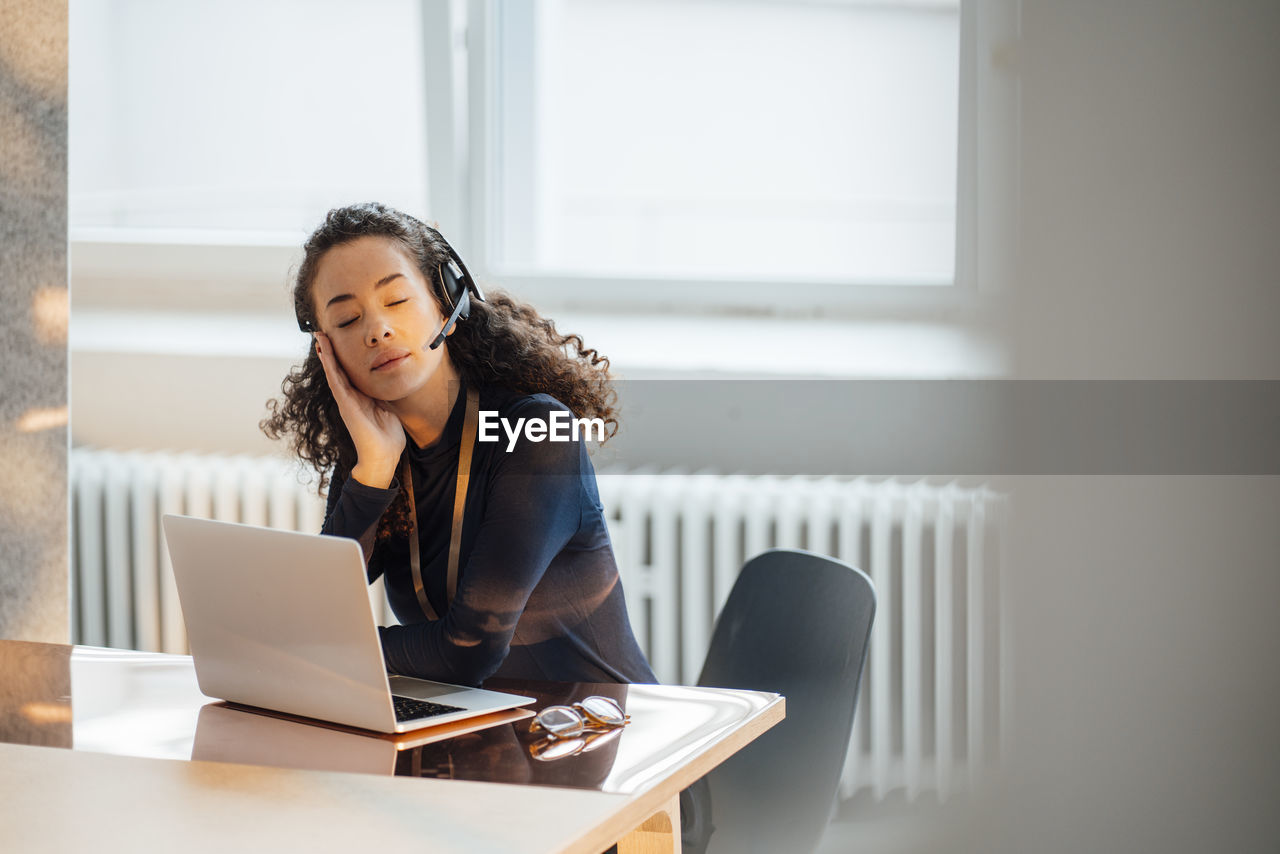 Young businesswoman wearing headset sitting with eyes closed at desk in office