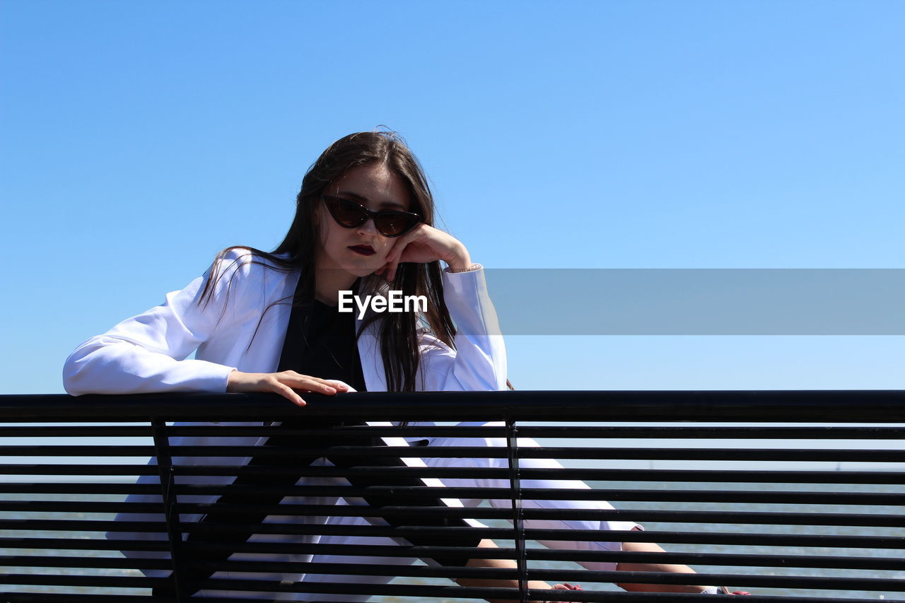 Confident girl sitting by railing against clear blue sky