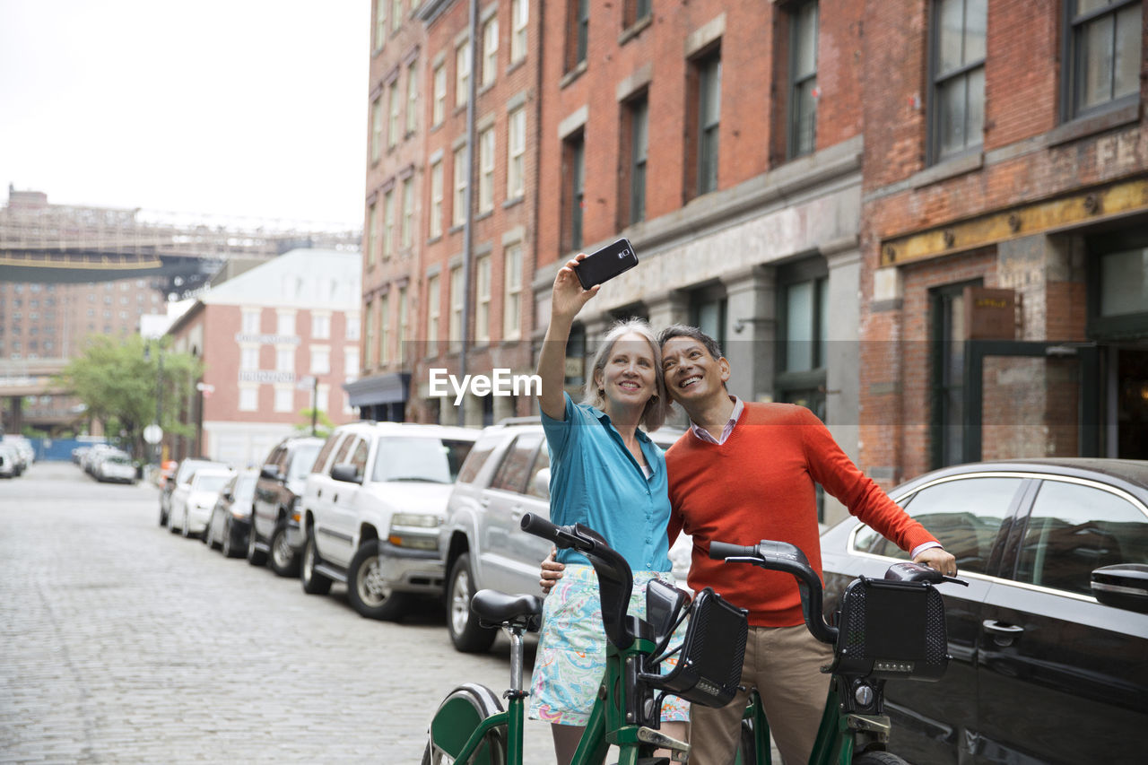 Happy mature couple taking selfie while standing with bicycles on city street