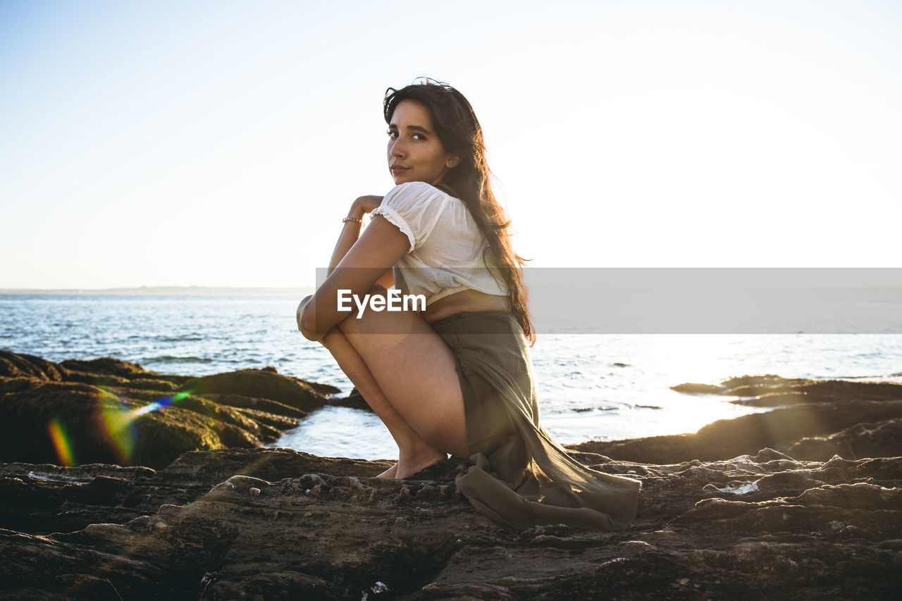 Young latina woman sitting by the ocean at golden hour in summertime