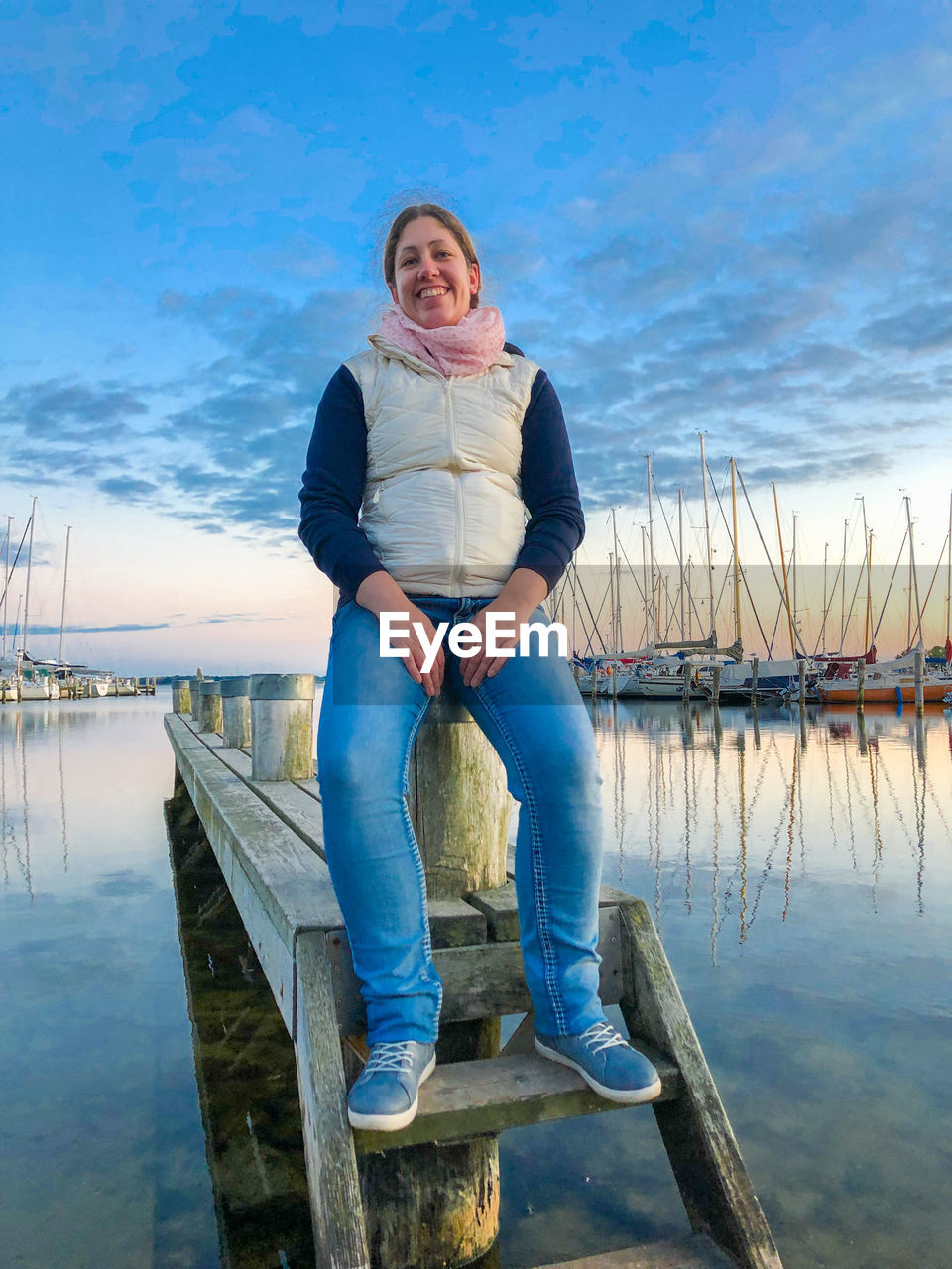 water, one person, full length, adult, sky, blue, nature, women, looking at camera, portrait, casual clothing, sea, smiling, front view, sitting, reflection, jeans, emotion, lifestyles, happiness, vacation, leisure activity, pier, day, cloud, clothing, footwear, wood, relaxation, female, young adult, person, outdoors, tranquility, beauty in nature, standing, nautical vessel, blond hair, jetty