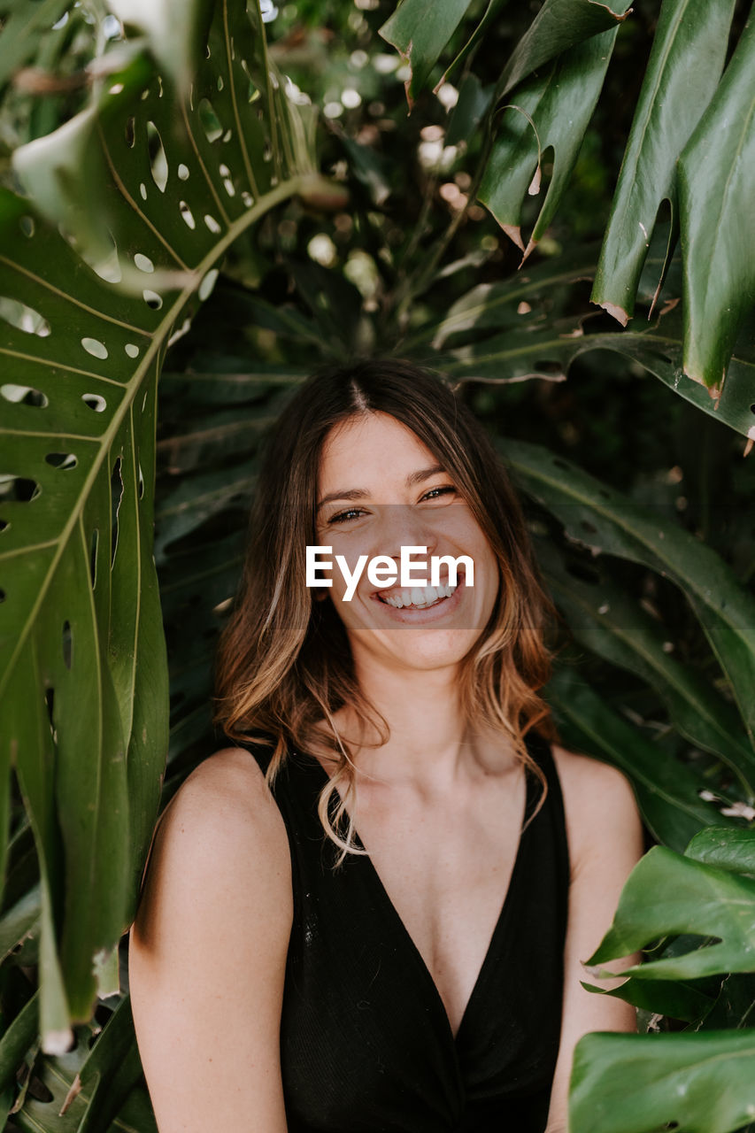 Satisfied female surrounded by green monstera plants looking at camera while standing in exotic garden in summer