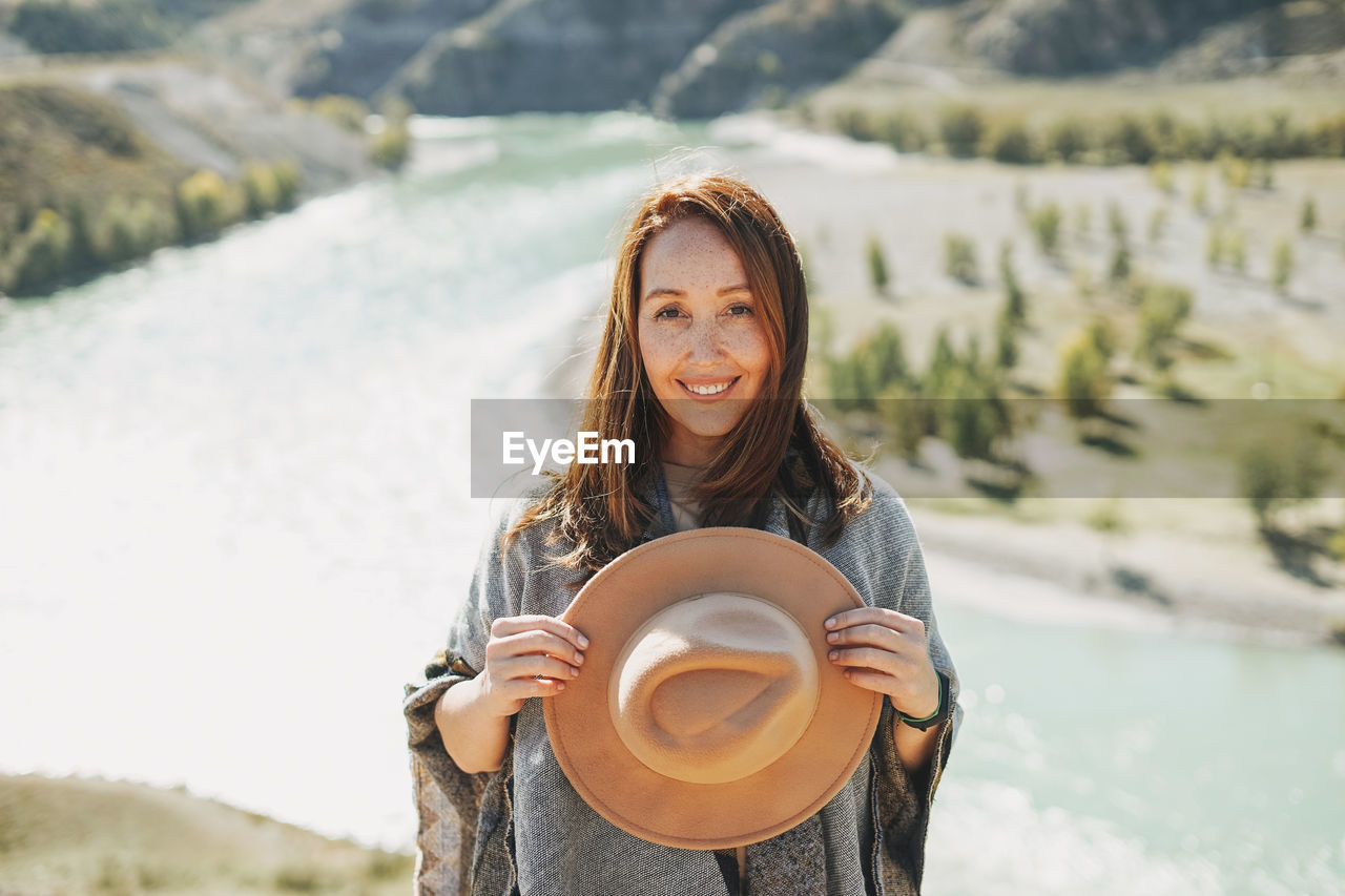 Carefree brunette young woman traveler in poncho with felt hat on background of mountain river