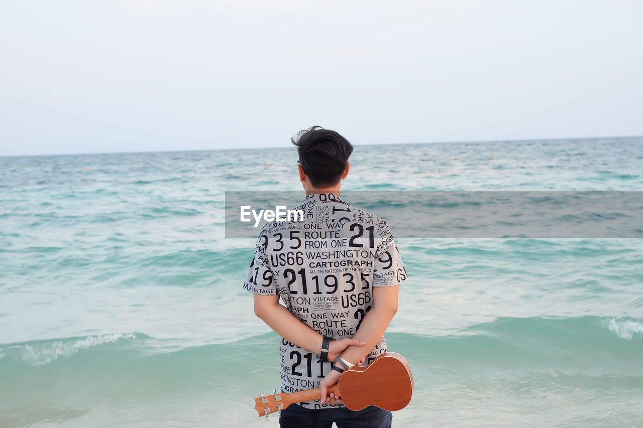 Rear view of young man holding violin while standing by sea against sky