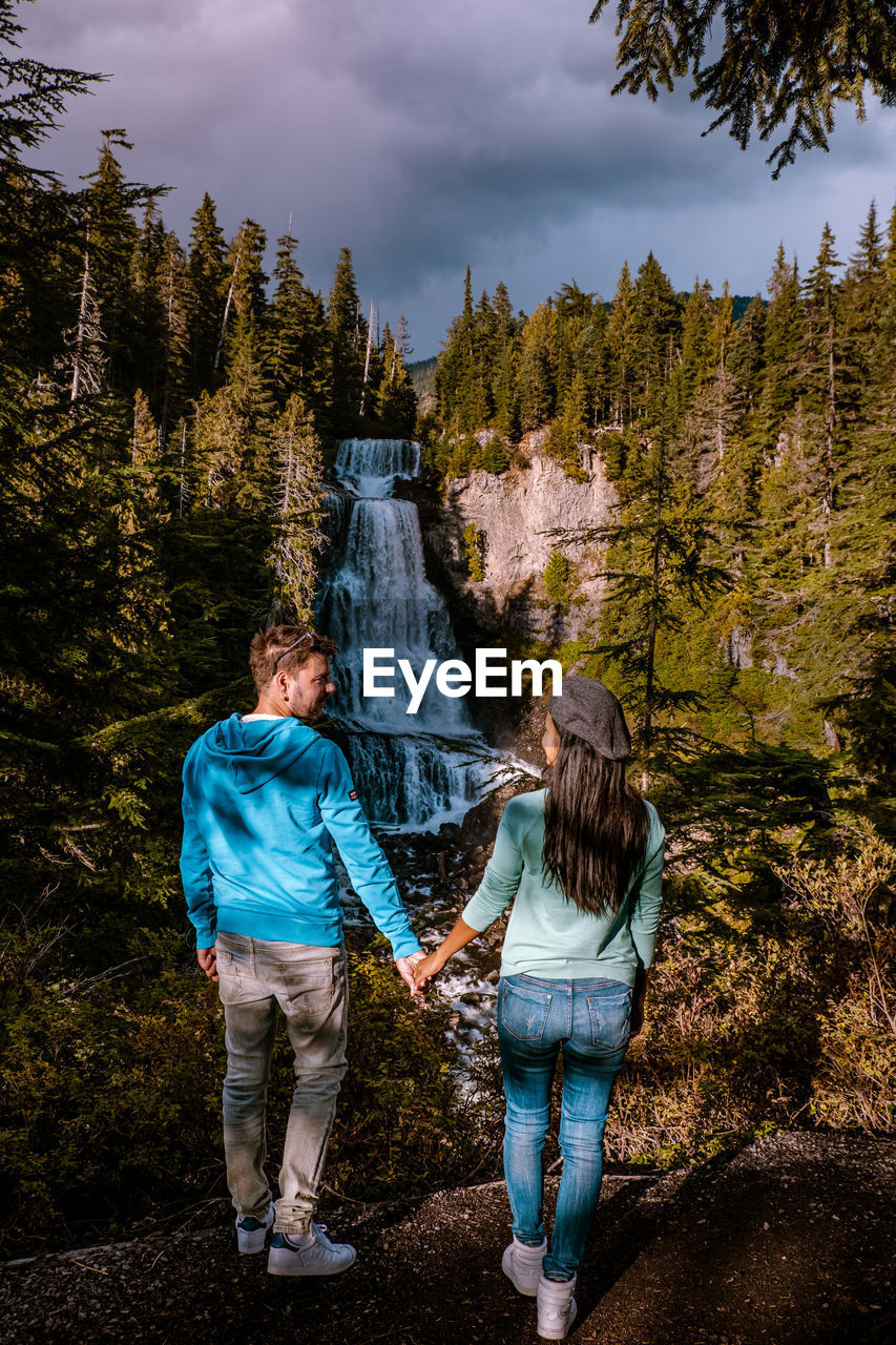 Rear view of people standing against waterfall and trees in forest