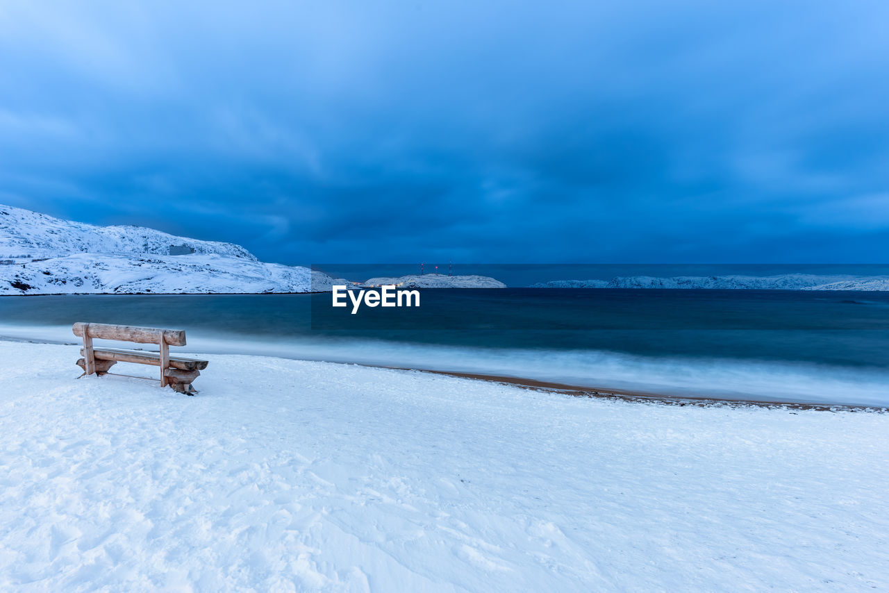 Lonely wooden bench overlooking the sea on a snow covered beach at dusk. long exposure.