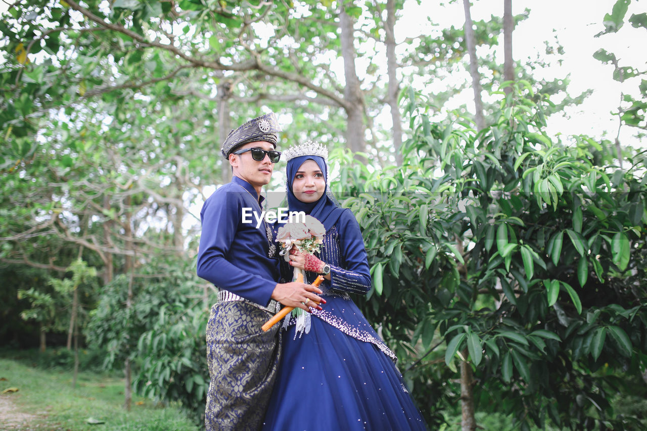 Bride and groom holding flower bouquet while standing by tree