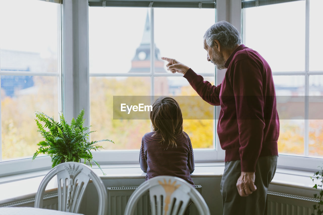 Side view of senior man showing something to great grandson through window at home