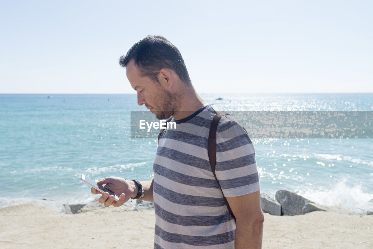 Side view of man using phone while standing at beach against clear sky