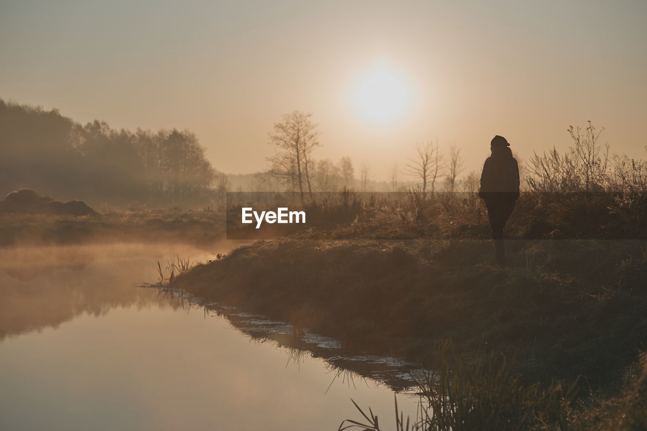 Rear view of silhouette man standing by lake against sky at sunrise