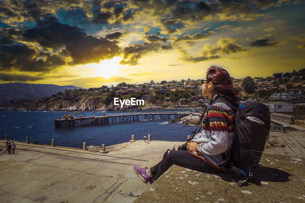 REAR VIEW OF WOMAN SITTING ON SHORE AGAINST SKY