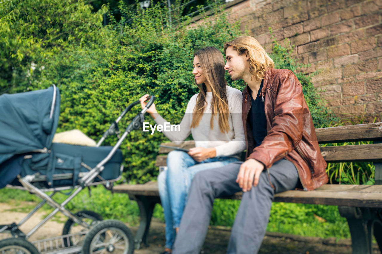 Portrait of smiling couple with baby stroller sitting on bench in park