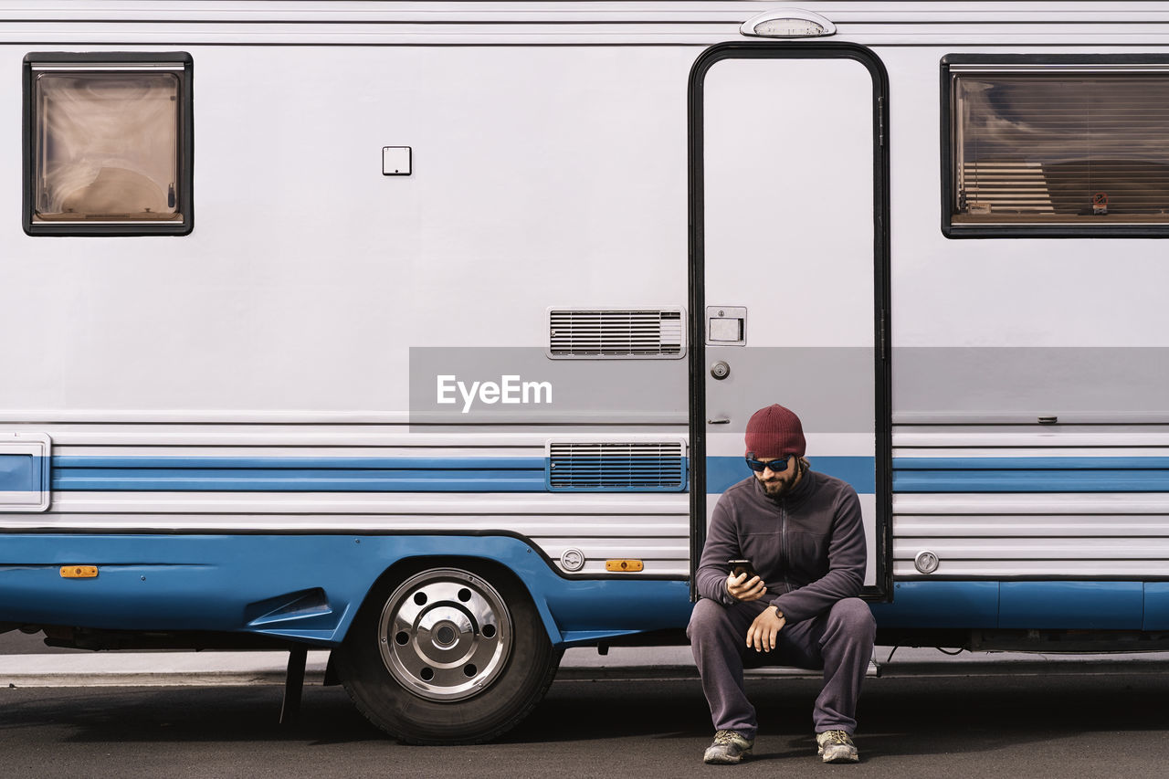 Full body of young bearded male traveler in casual clothes and sunglasses sitting on step of camper van and messaging on smartphone