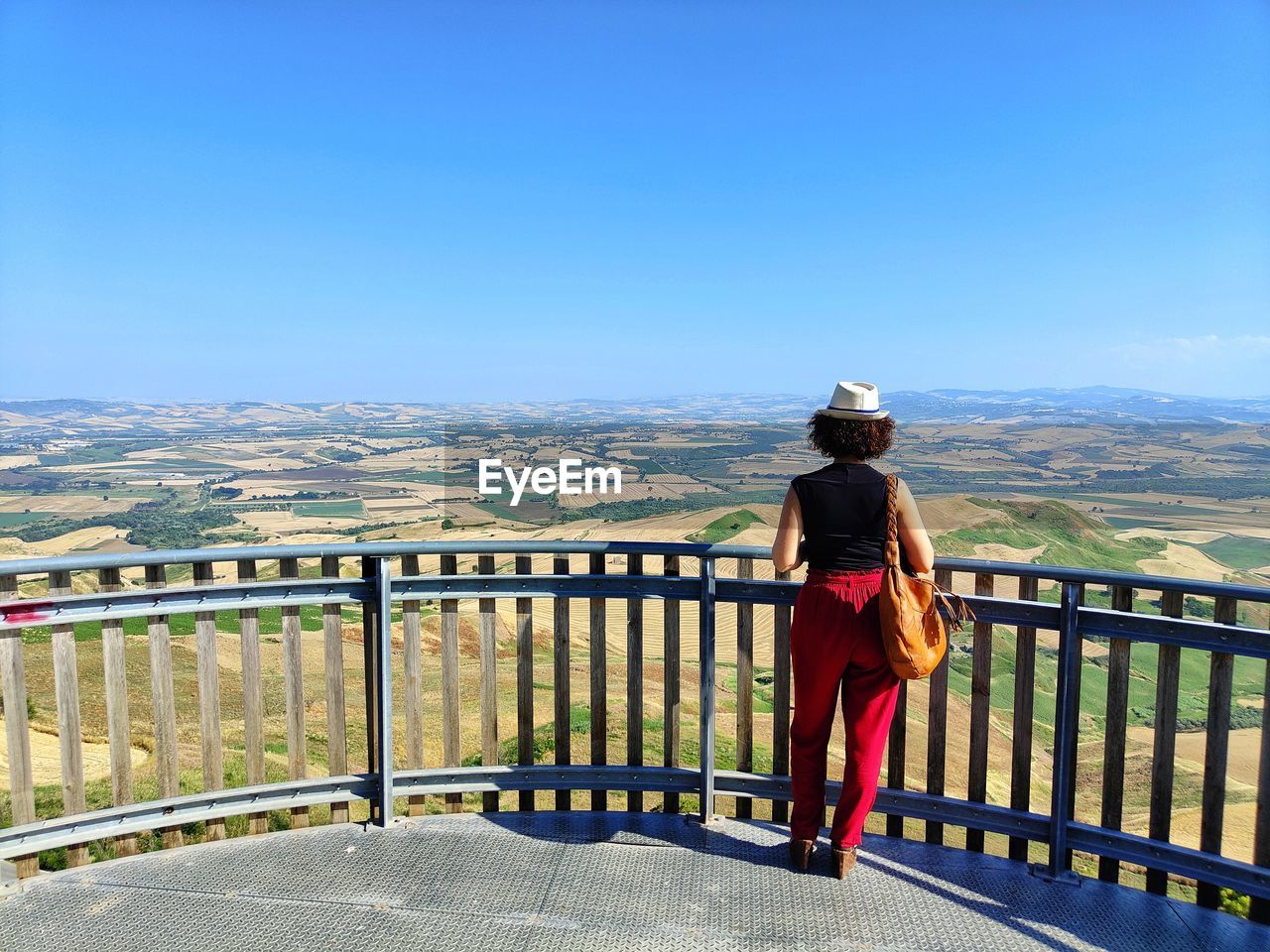 railing, vacation, full length, one person, rear view, sky, nature, mountain, adult, looking at view, architecture, day, lifestyles, clothing, leisure activity, scenics - nature, standing, beauty in nature, casual clothing, clear sky, women, travel, sunlight, landscape, fence, backpack, person, built structure, blue, sunny, copy space, outdoors, travel destinations, city, walking, mountain range, tourism, hat, transportation, men, water, observation point, footwear, activity, tranquil scene, young adult, tranquility, road