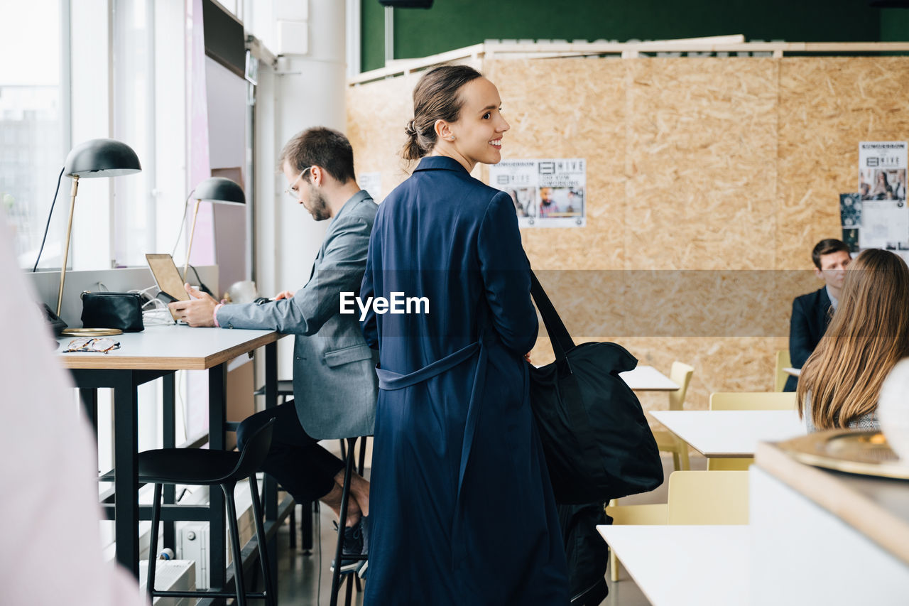 Smiling businesswoman with bag looking away while standing amidst colleagues in coworking space