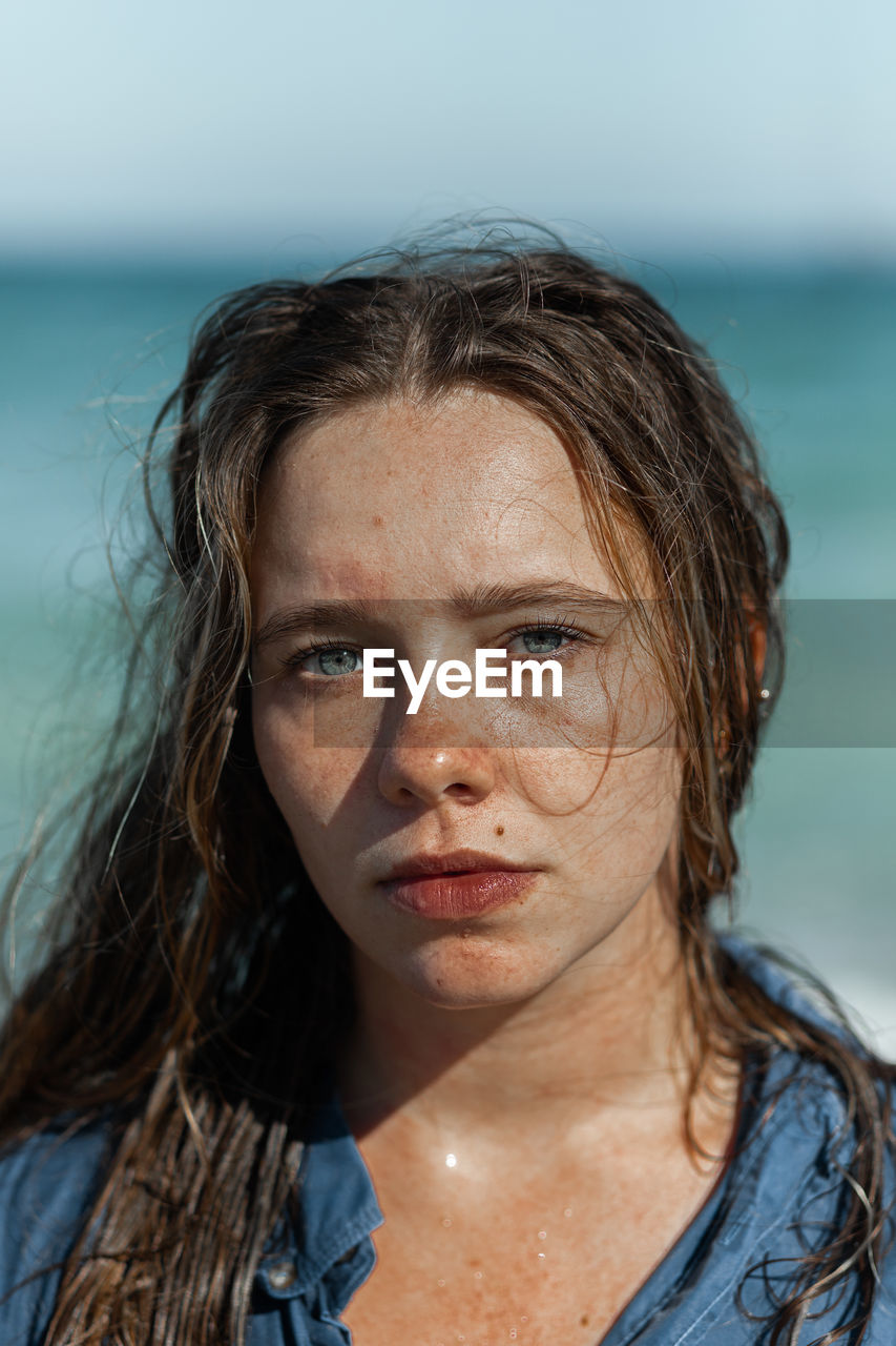 Female in wet shirt and with wet hair standing looking at camera on beach near sea while enjoying summer day