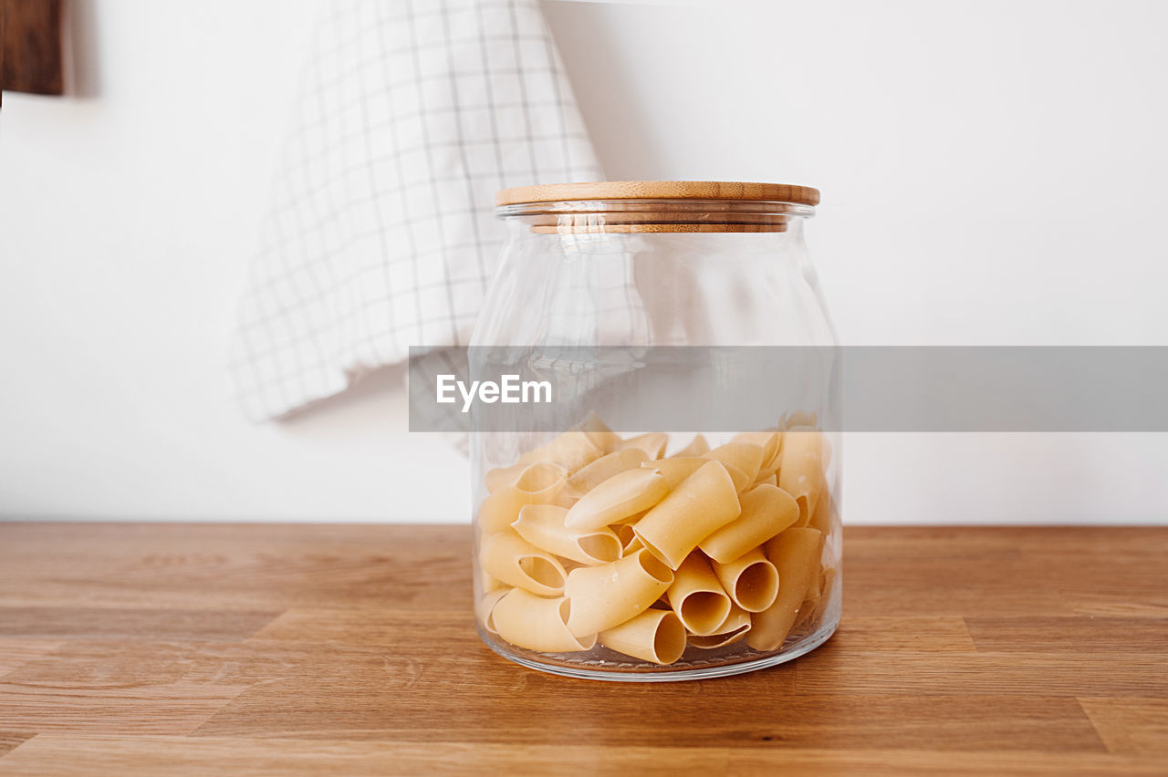 Dry raw large macaroni from a glass large jar with a wooden eco lid on a wooden countertop 
