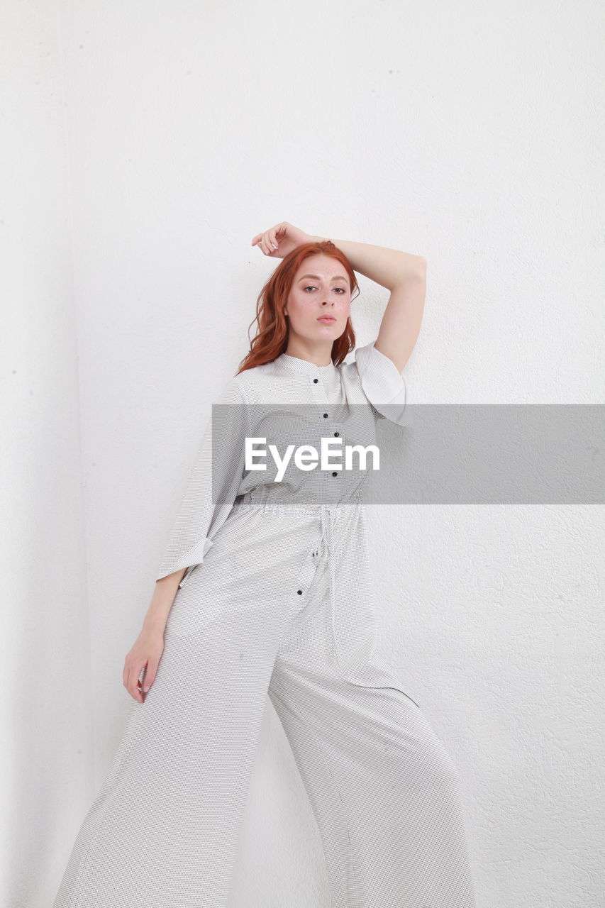 Cropped fashion catalog style portrait of woman in jumpsuit standing on mirror over white wall