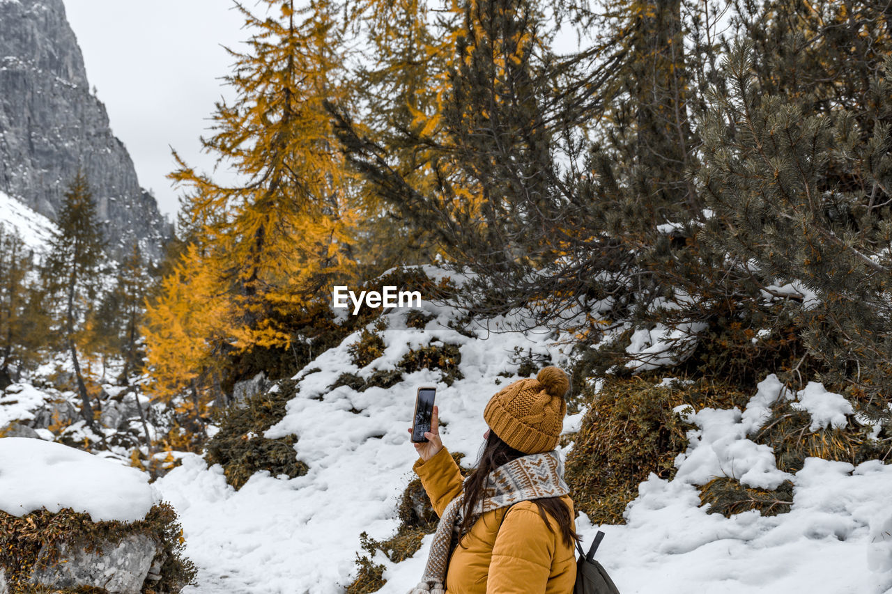 Side view of young woman hiking on snowy path, taking photos of mountains